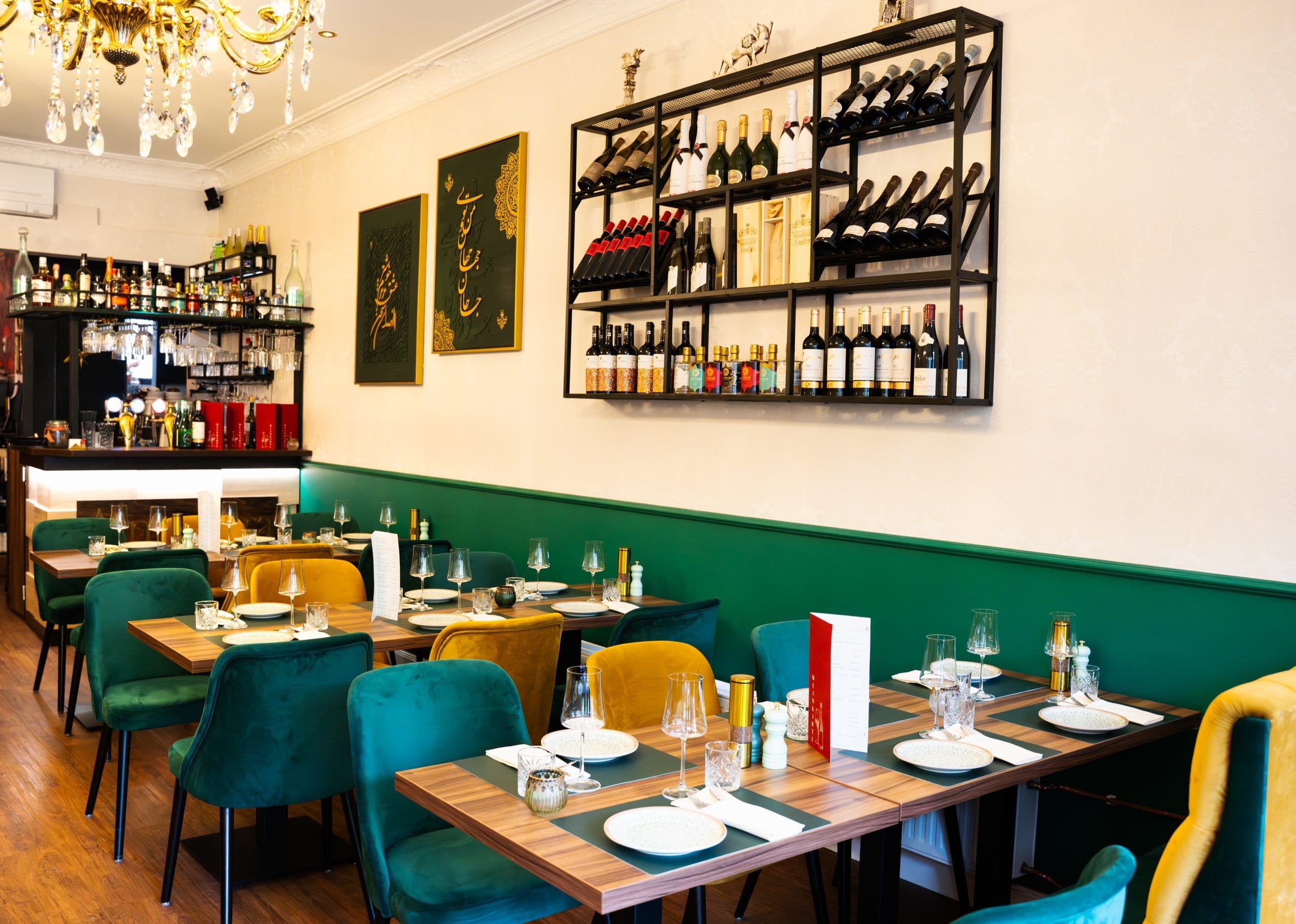 Gastraum mit eingedeckten Holztischen, petrolfarbenen und senfgelben Samtstühlen, Weinregal und Bar im Hintergrund.